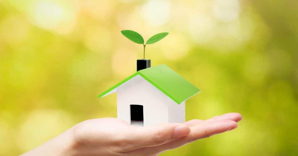 A close-up of a person holding a model home in their hand. The home has a green roof with a sprout coming through the chimney.