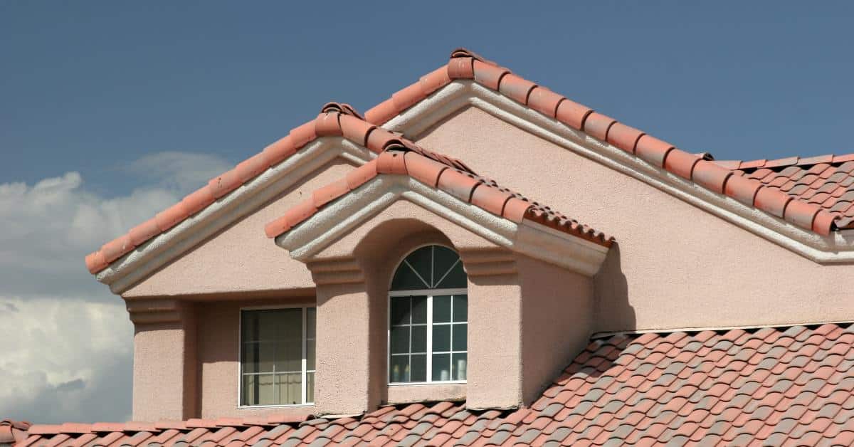 A home with a red clay tile roof, stucco walls, and multiple gables beneath a clear blue sky on a sunny day.