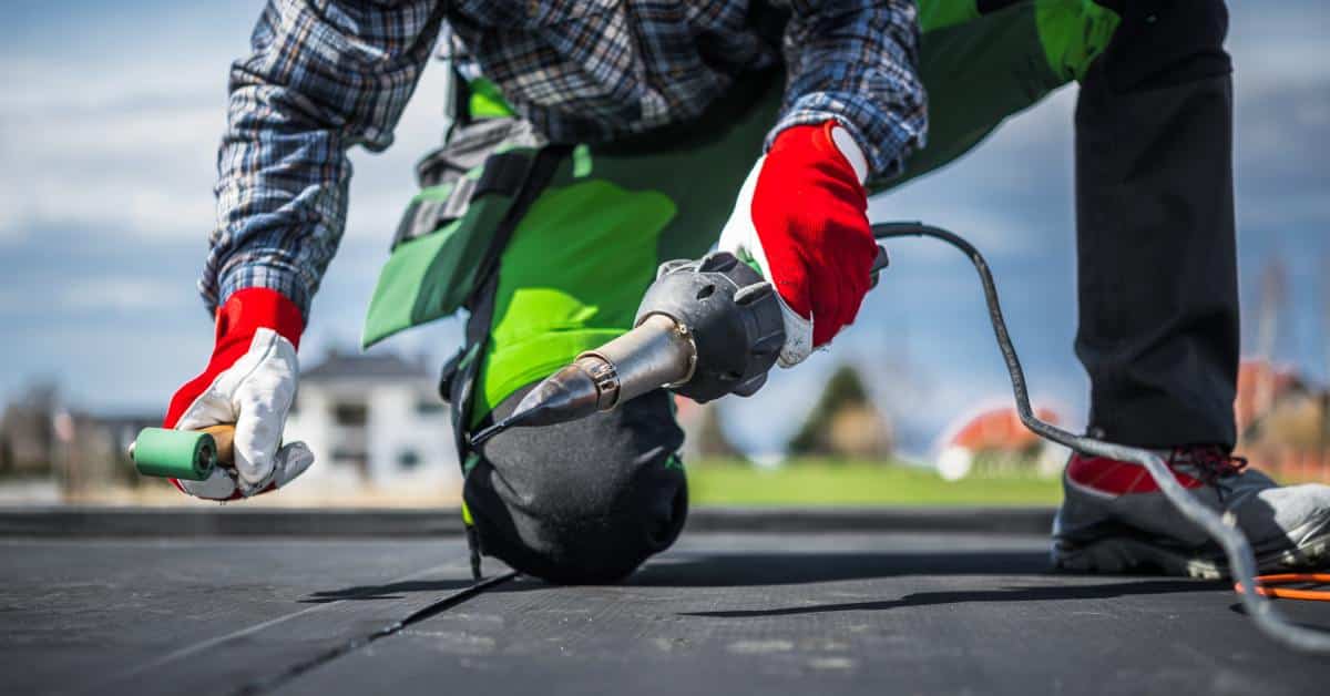 A roofer in red gloves kneels on a flat roof, using a hot air gun and a roller to install black EPDM roofing material.