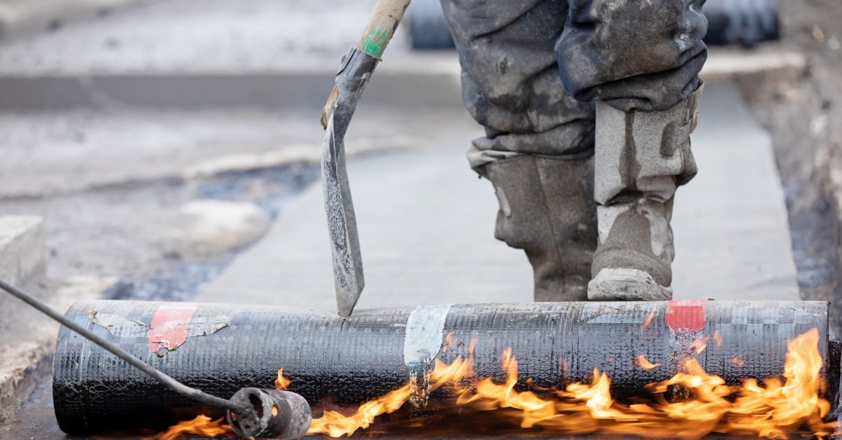 A roofer stands behind a roll of black modified bitumen roofing membrane as its base is heated with a flaming torch.