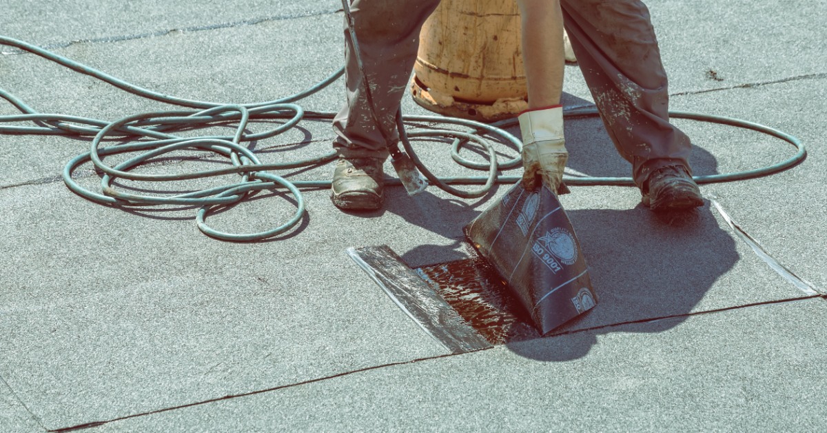 A roofer uses a torch to patch a section of a modified bitumen roof. He holds up a membrane panel to heat the adhering layer.