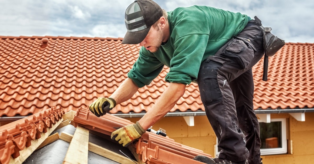 A roofer in gloves bends over as he installs terracotta tiles on a sloped roof under gray clouds.