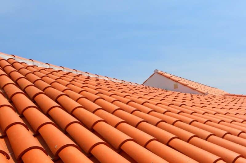 A wide view of a sunlit terracotta tile roof, showing rows of curved tiles leading toward a clear blue sky.