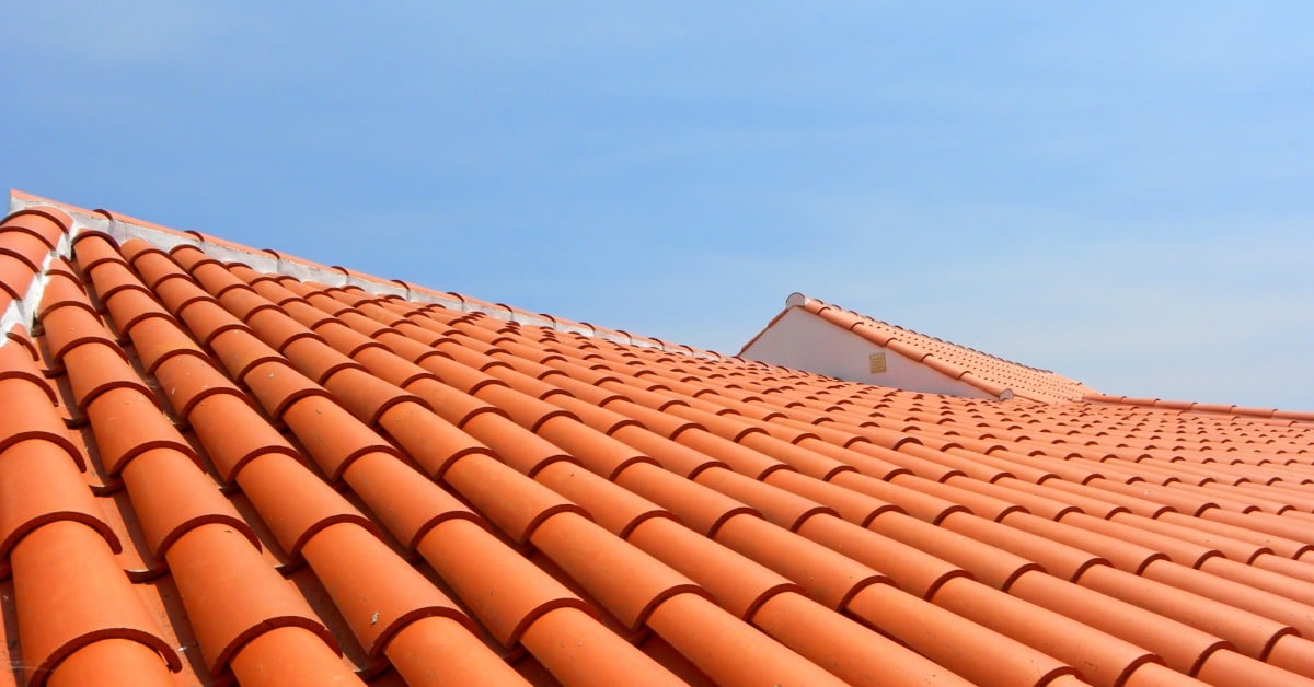 A wide view of a sunlit terracotta tile roof, showing rows of curved tiles leading toward a clear blue sky.