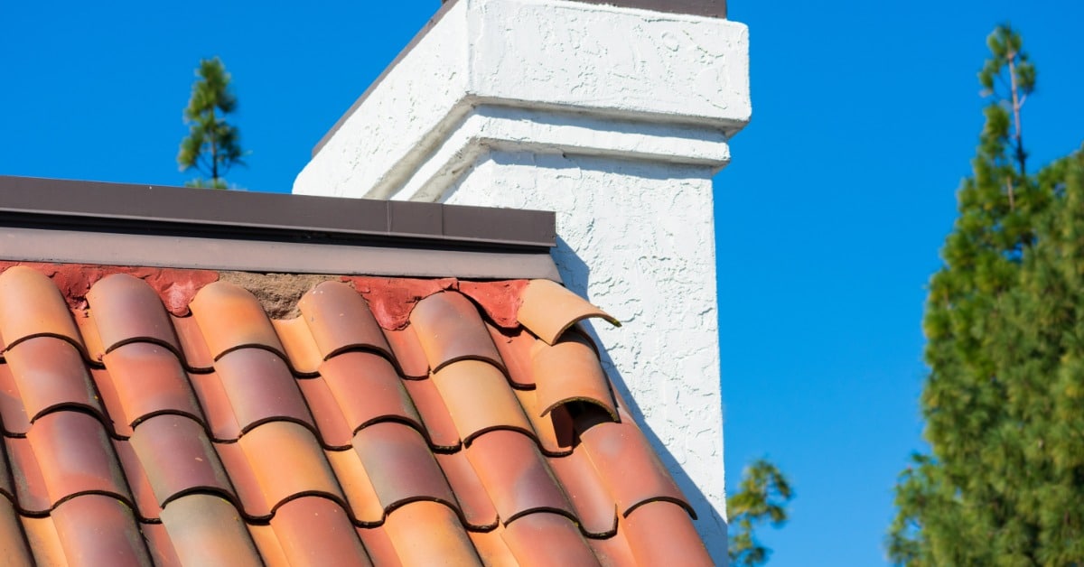 A white chimney rises from a terracotta tile roof with two dislodged tiles, set against a bright blue sky.