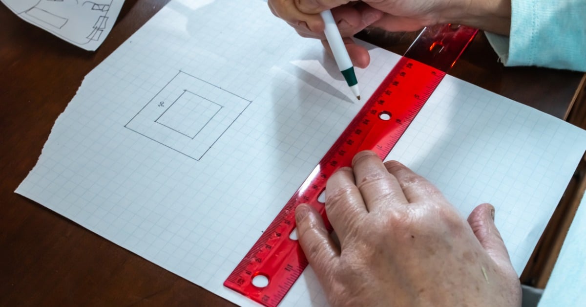 A close-up of a person using a pen and a bright red plastic ruler to make area calculations on grid paper.