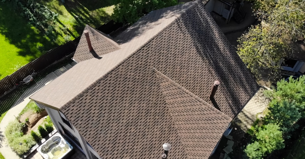An aerial view of a large shingle gabled roof with a dormer on either side. The home is surrounded by trees and yard space.