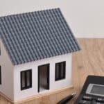 A model house with a rectangular gabled roof sits beside a calculator and pen on a wooden table against a white wall.