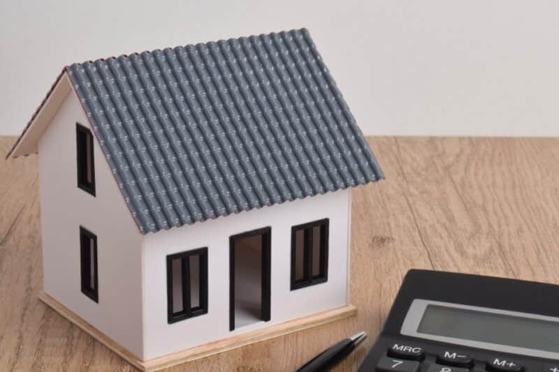 A model house with a rectangular gabled roof sits beside a calculator and pen on a wooden table against a white wall.