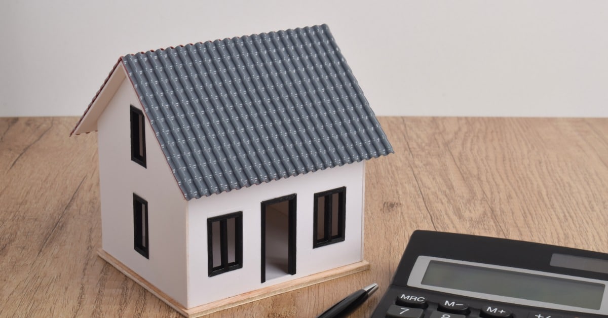 A model house with a rectangular gabled roof sits beside a calculator and pen on a wooden table against a white wall.