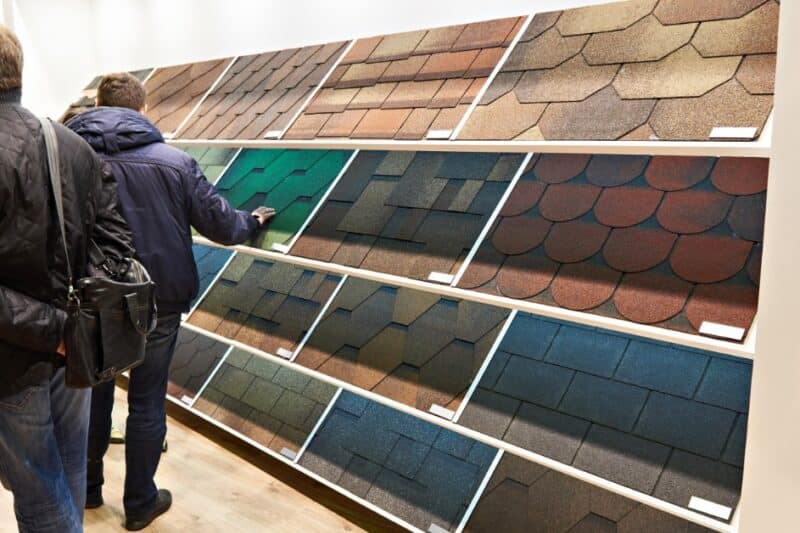 Two customers browse a showroom wall of roofing shingle sample panels of varying colors and shapes on display.