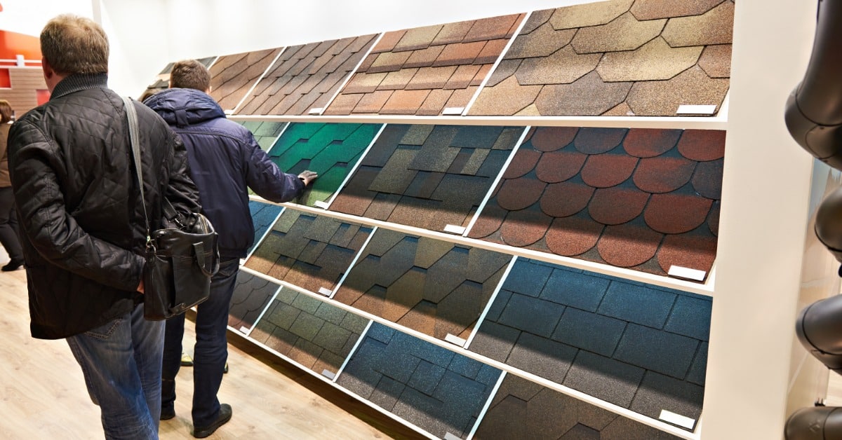 Two customers browse a showroom wall of roofing shingle sample panels of varying colors and shapes on display.