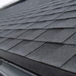 a dark gray shingled roof with a black rain gutter and downspout running along the lower edge near a blurred house.