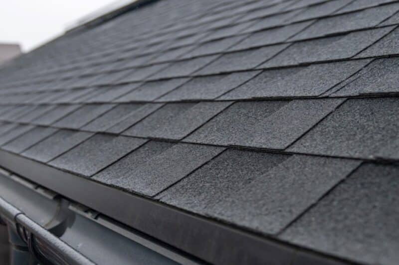 a dark gray shingled roof with a black rain gutter and downspout running along the lower edge near a blurred house.