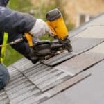 A person in a reflective vest uses a yellow nail gun to attach gray shingles across a sloped roof surface.
