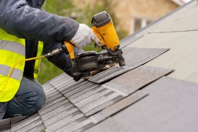 A person in a reflective vest uses a yellow nail gun to attach gray shingles across a sloped roof surface.