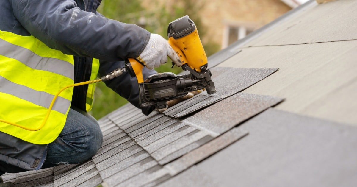 A person in a reflective vest uses a yellow nail gun to attach gray shingles across a sloped roof surface.