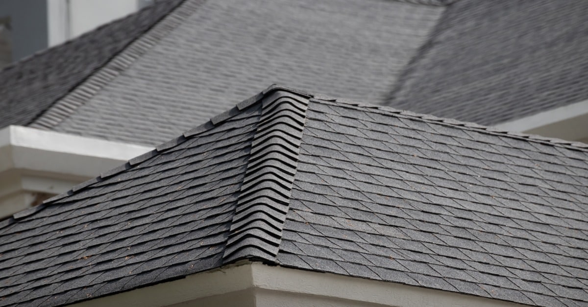A roof has gray asphalt shingles across several slopes, with layered ridges and a light building exterior below.