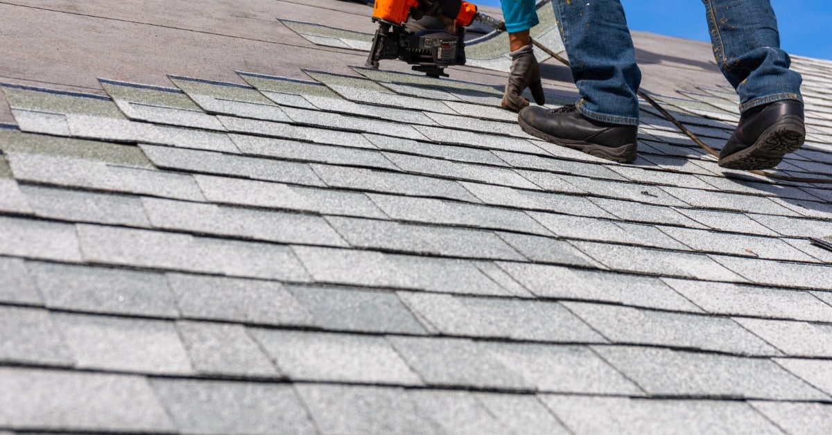 A person uses an orange nail gun while standing on gray roof shingles, with a blue sky above the roofline.
