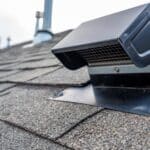 A black static roof vent installed on gray asphalt shingles, with roof pipes blurred in the background.