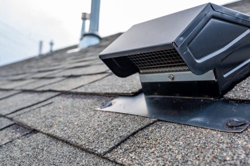 A black static roof vent installed on gray asphalt shingles, with roof pipes blurred in the background.
