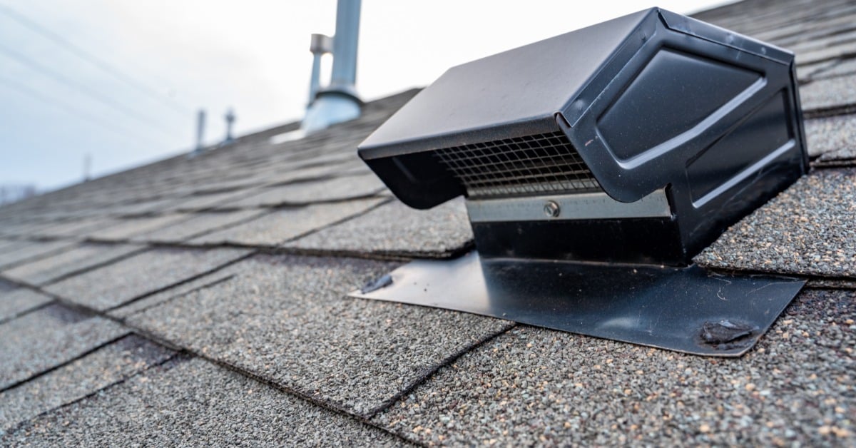 A black static roof vent installed on gray asphalt shingles, with roof pipes blurred in the background.