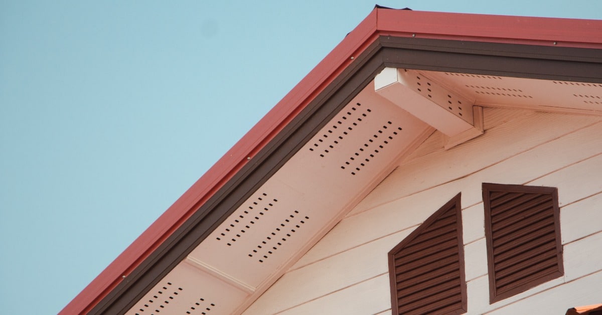 A house roof corner with red trim, beige soffit vent holes, brown shutters, and clear blue sky overhead.