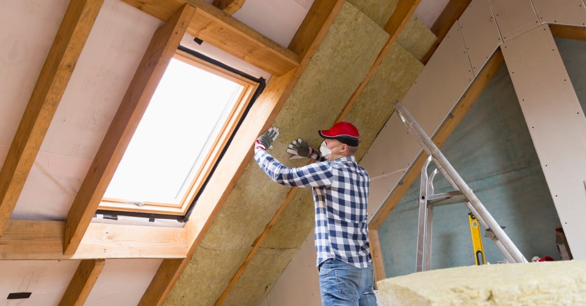 A person wearing gloves and a red cap installing yellow insulation panels between wooden attic beams.