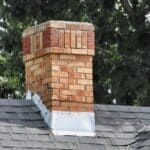 A brick chimney rises from a gray shingled roof with metal flashing and leafy green trees in the background.