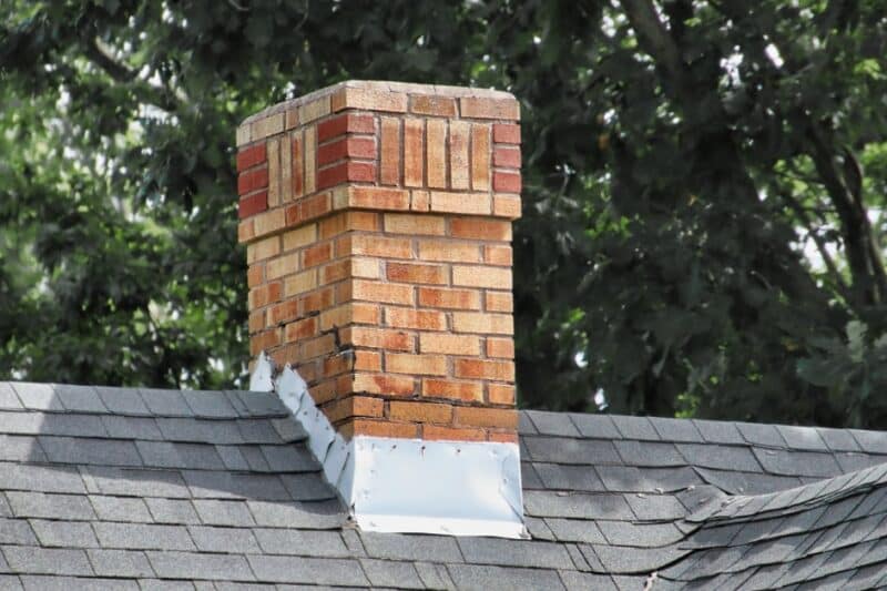 A brick chimney rises from a gray shingled roof with metal flashing and leafy green trees in the background.