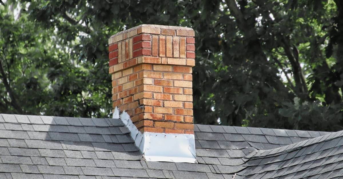 A brick chimney rises from a gray shingled roof with metal flashing and leafy green trees in the background.