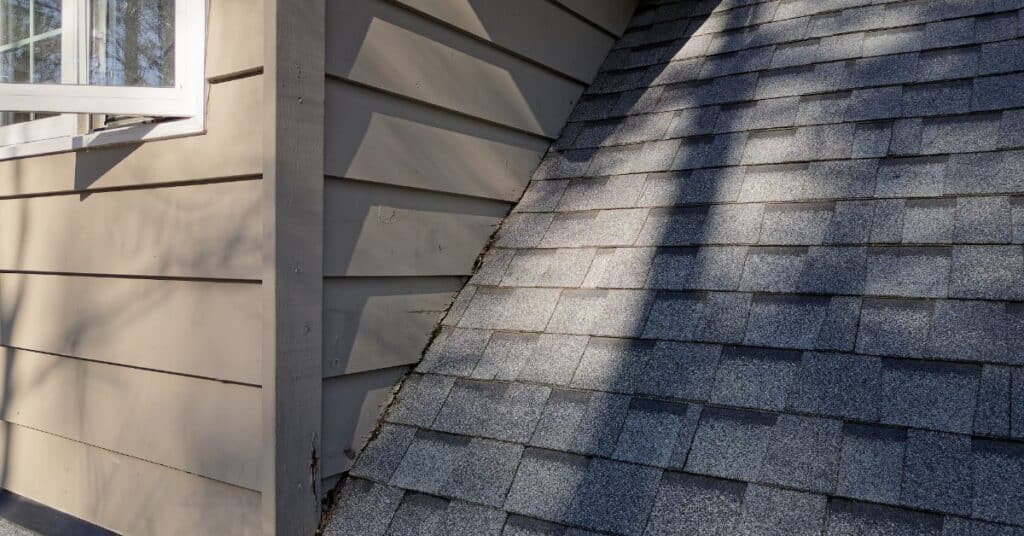 A damaged section of beige wood siding meets a gray shingled roof below a white window with shadows across the surface.