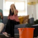 A red bucket catching water drops on a wood floor, with two blurred people seated on a couch in the background.