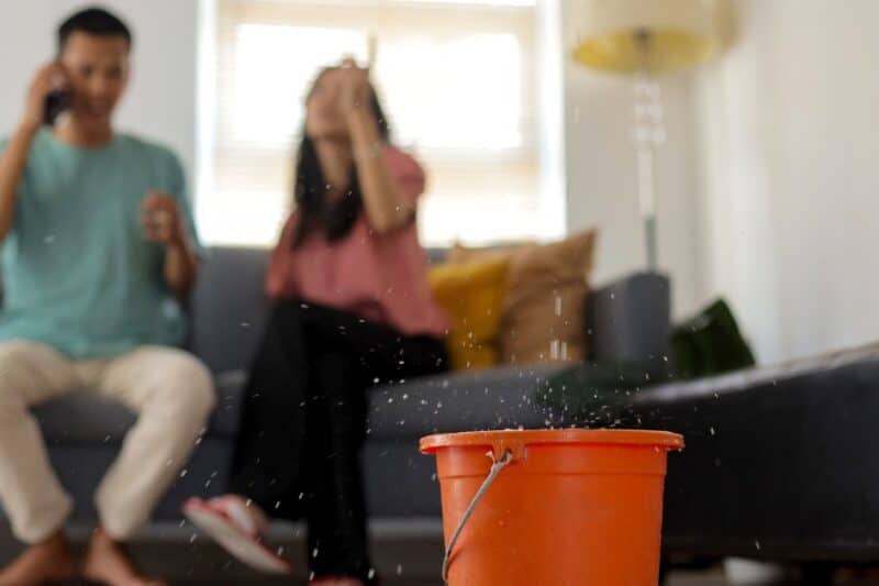 A red bucket catching water drops on a wood floor, with two blurred people seated on a couch in the background.
