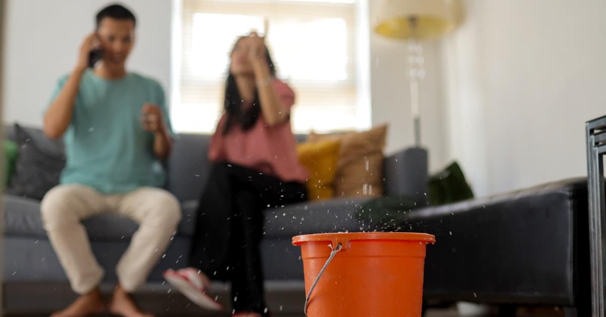 A red bucket catching water drops on a wood floor, with two blurred people seated on a couch in the background.