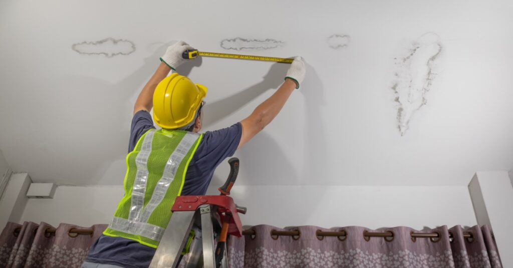 A worker wearing a yellow hard hat measures ceiling stains while standing on a ladder above patterned curtains.