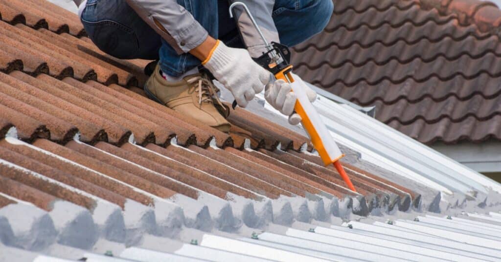 A worker wearing gloves applies sealant from a caulking gun along red roof tiles beside white metal panels.
