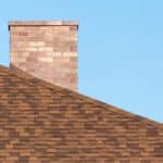 A brick chimney rising above brown shingles under a clear blue sky, with the roof sloping across the lower area.