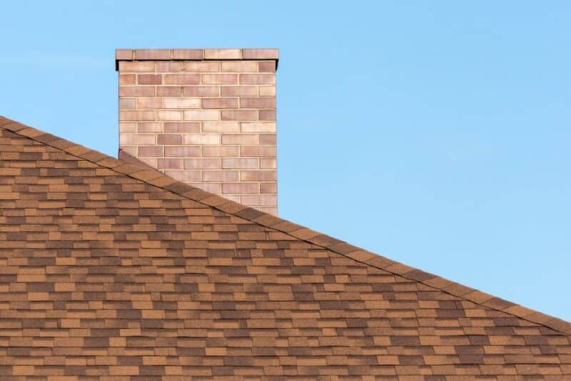 A brick chimney rising above brown shingles under a clear blue sky, with the roof sloping across the lower area.