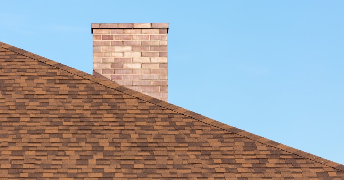 A brick chimney rising above brown shingles under a clear blue sky, with the roof sloping across the lower area.