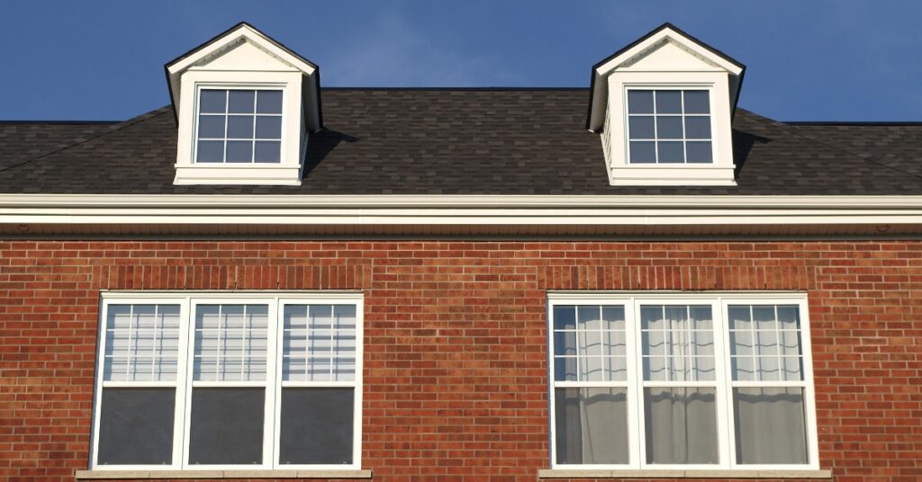 A red brick house beneath a dark shingle roof has two dormer windows and white-trimmed lower windows under blue sky.