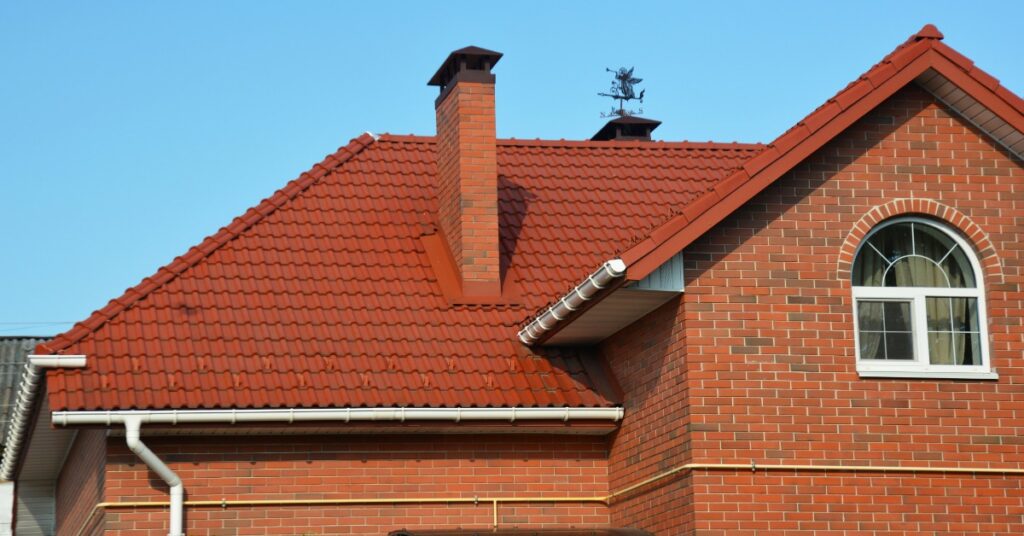 A red tile roof covers a brick house with a chimney, white gutters, an arched window, and blue sky above.