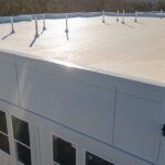 A white flat roof on a large building with short pipe vents, windows, trees, and clear sky in the background.
