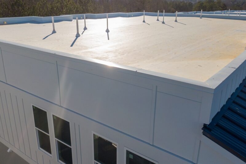 A white flat roof on a large building with short pipe vents, windows, trees, and clear sky in the background.