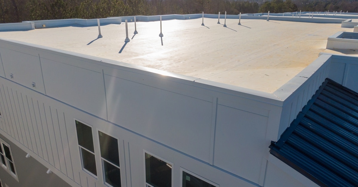 A white flat roof on a large building with short pipe vents, windows, trees, and clear sky in the background.