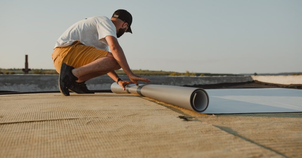 A man kneeling on a flat roof while rolling out a light gray PVC membrane across a textured surface under a clear sky.