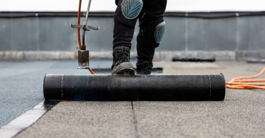 A person wearing knee pads and boots applying a black Bitumen roofing roll on a flat surface with a torch and orange hose.