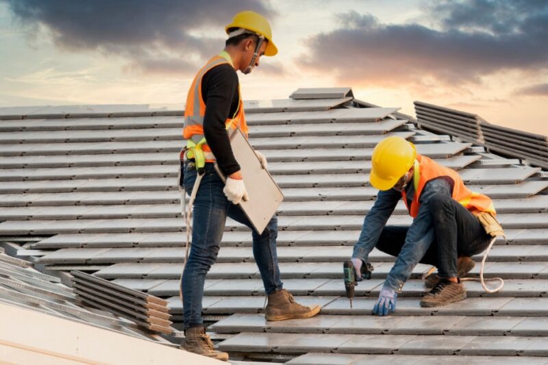 A pair of men in yellow hard hats and orange safety vests installing gray roof tiles with a cordless drill.