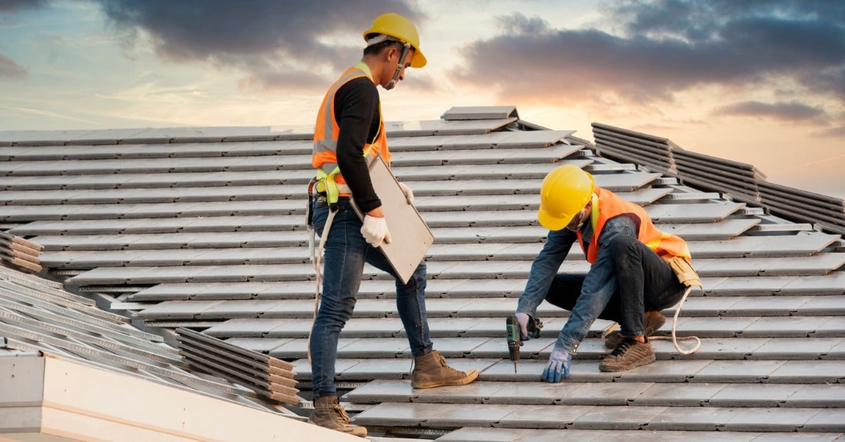 A pair of men in yellow hard hats and orange safety vests installing gray roof tiles with a cordless drill.