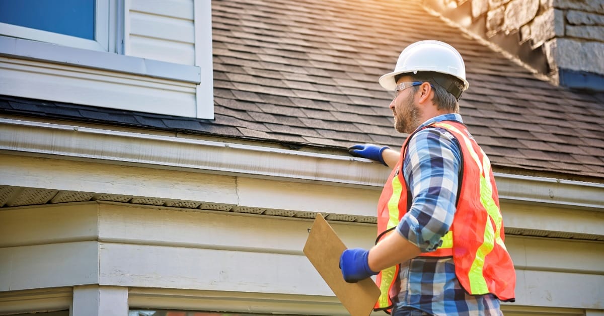 A man in a white hard hat and orange safety vest touching the roof gutter while holding a clipboard beside a house.